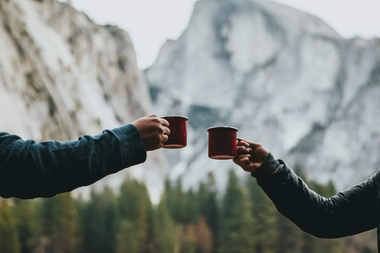 Two people clinking red mugs outdoors with snowy mountains behind them