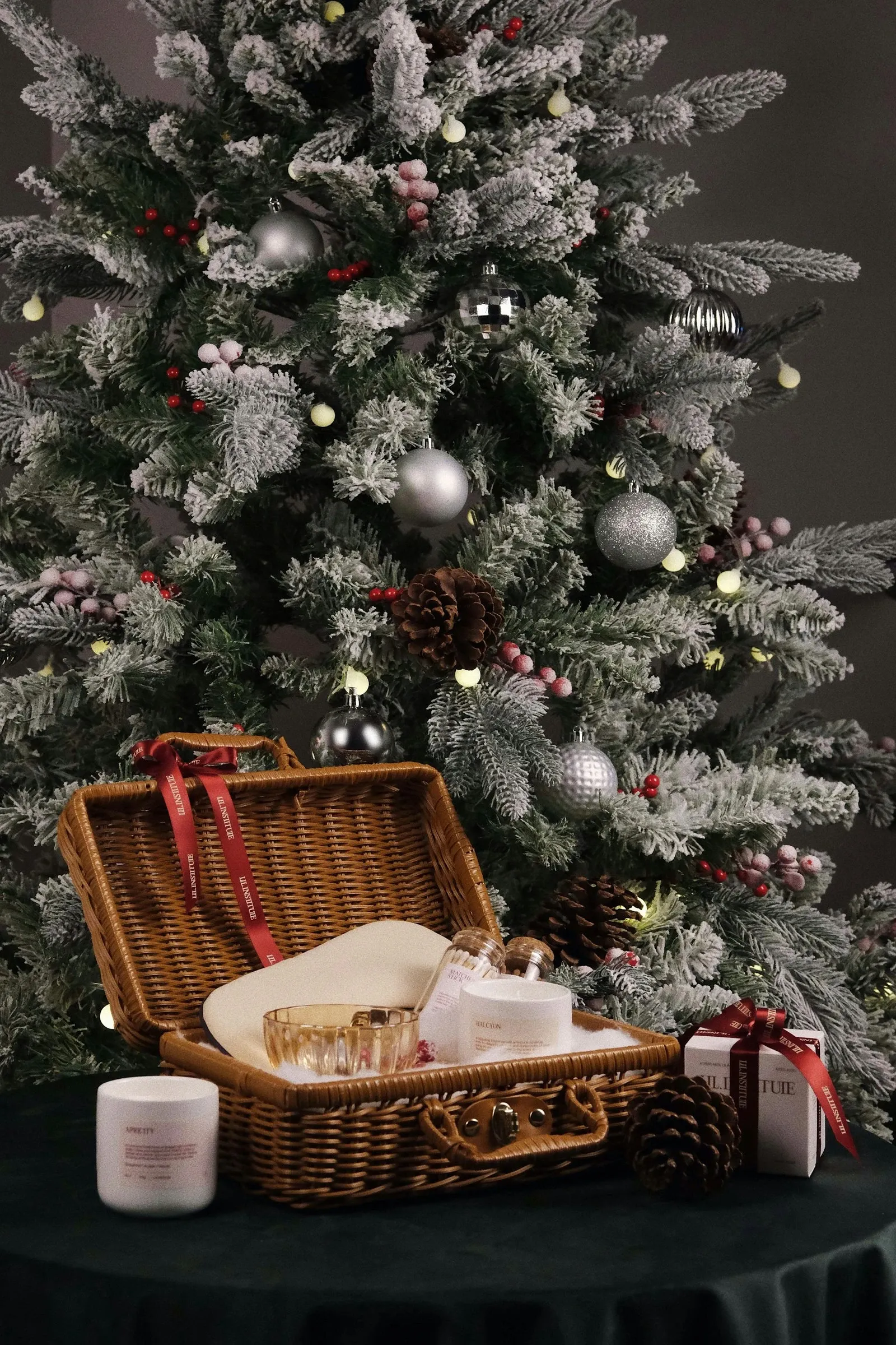 Wicker picnic basket of gifts with a red bow under a frosted Christmas tree hung with silver ornaments