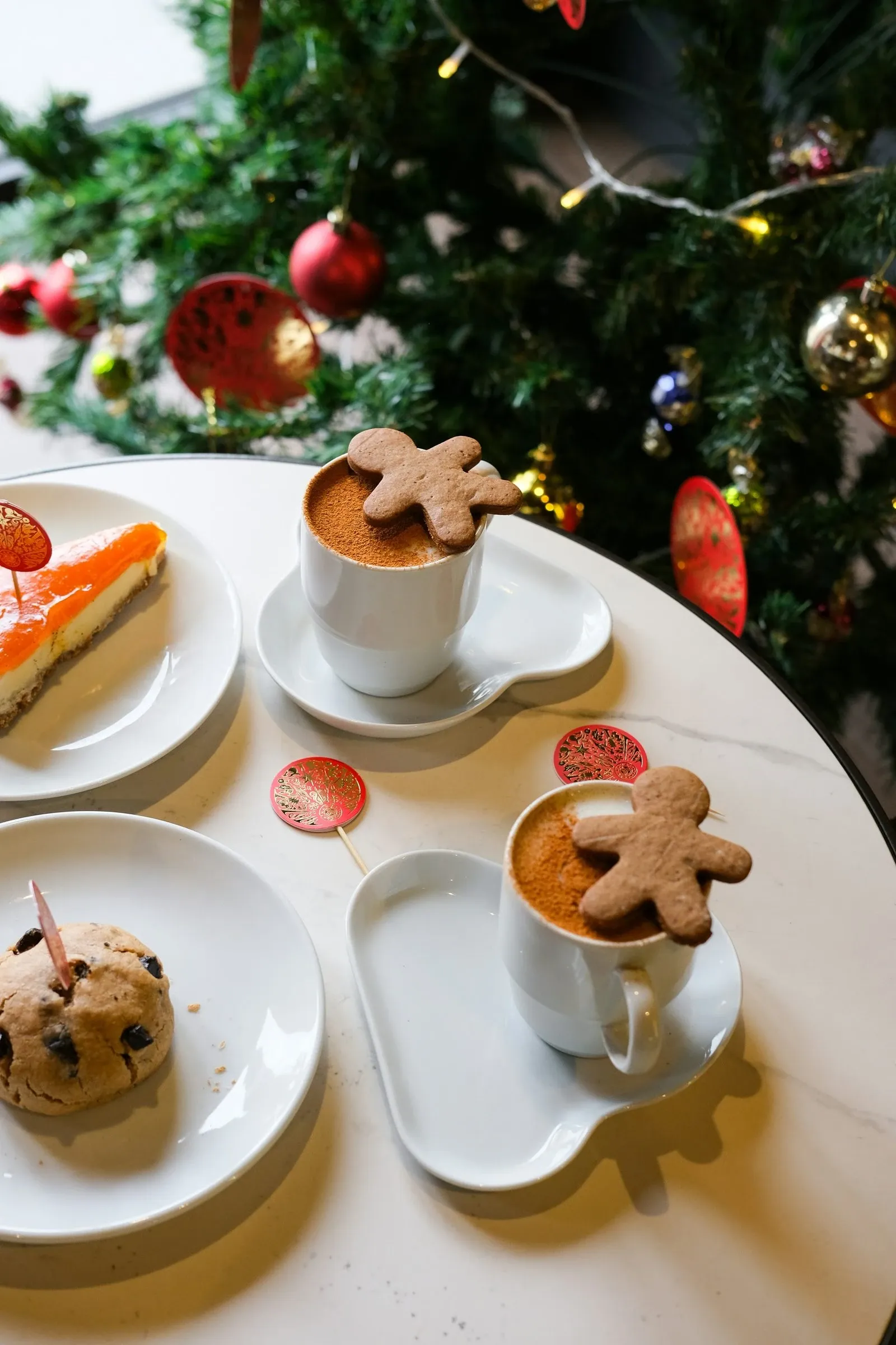 Two lattes with gingerbread man cookies on plates near a decorated Christmas tree