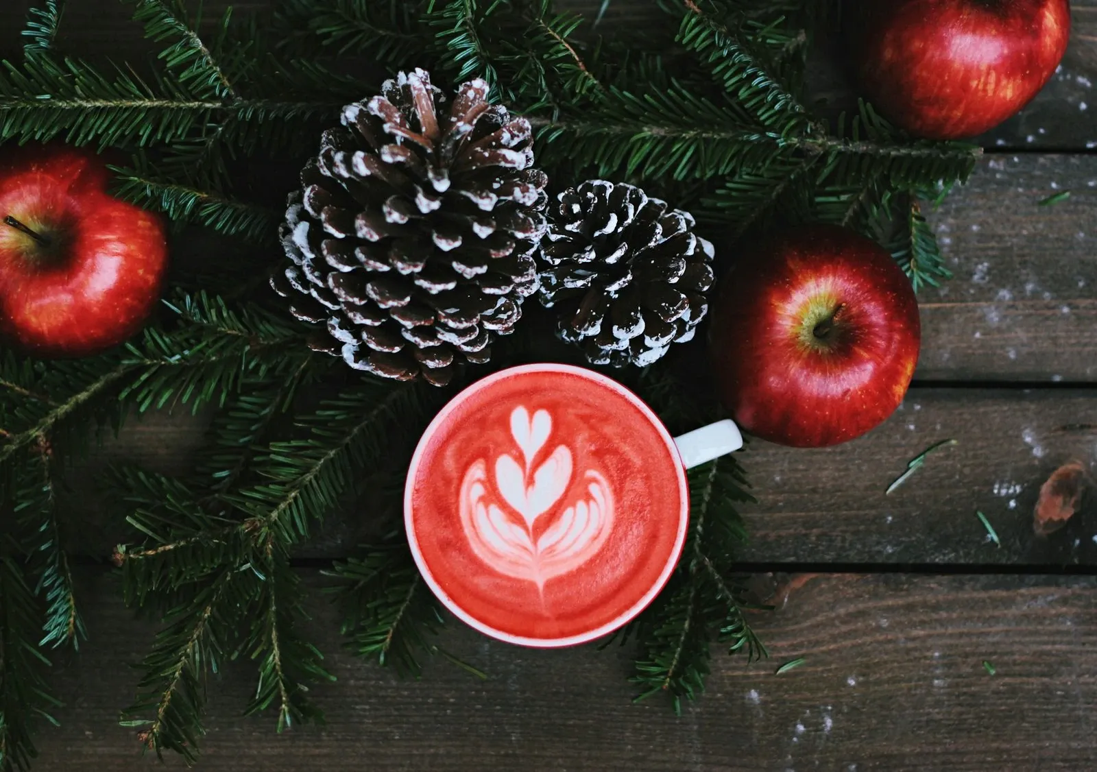 Red mug with heart latte art on a wood board surrounded by pinecones, red apples and pine sprigs