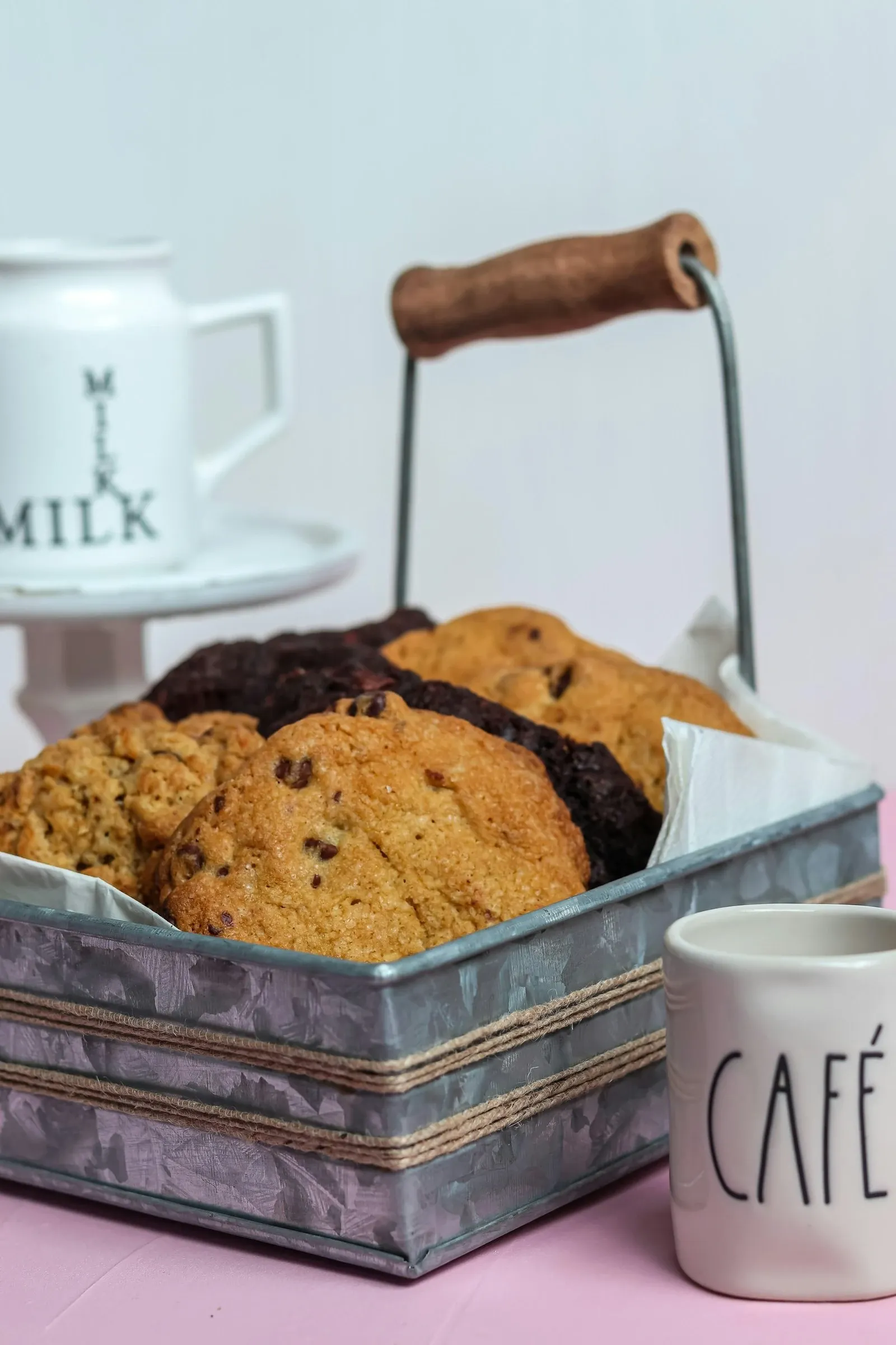 Chocolate chip cookies in a galvanized metal caddy with a wooden handle beside white cafe and milk mugs