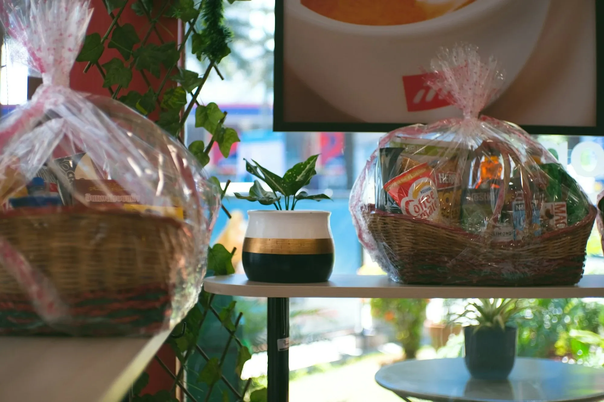 Two cellophane-wrapped gift baskets with colorful snacks displayed on cafe shelves near a window