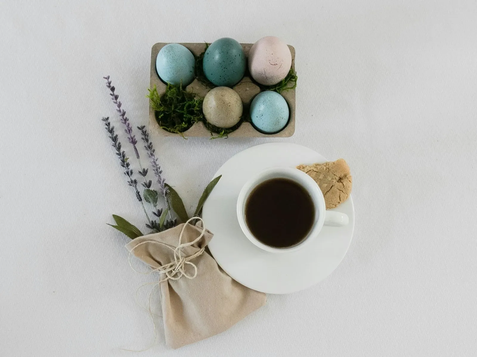 White coffee mug beside a plate of pastel eggs and a sprig of lavender on a white table