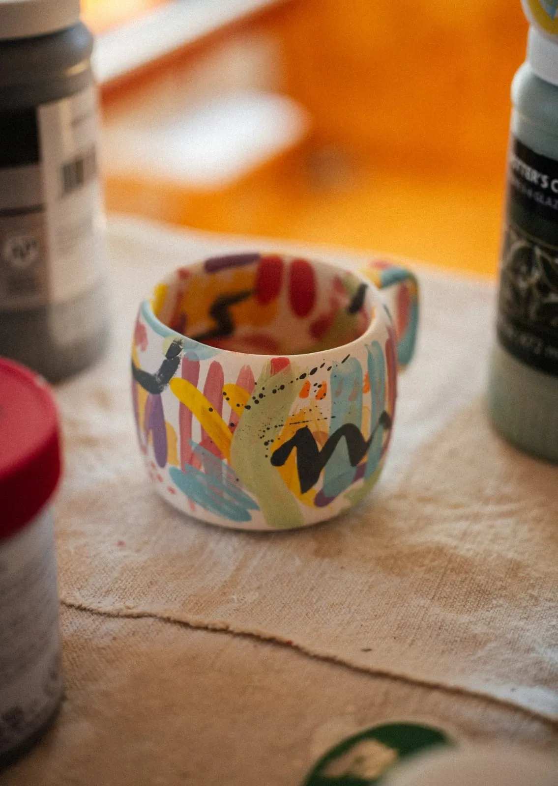 Ceramic mug hand-painted with colorful abstract shapes sitting among paint jars
