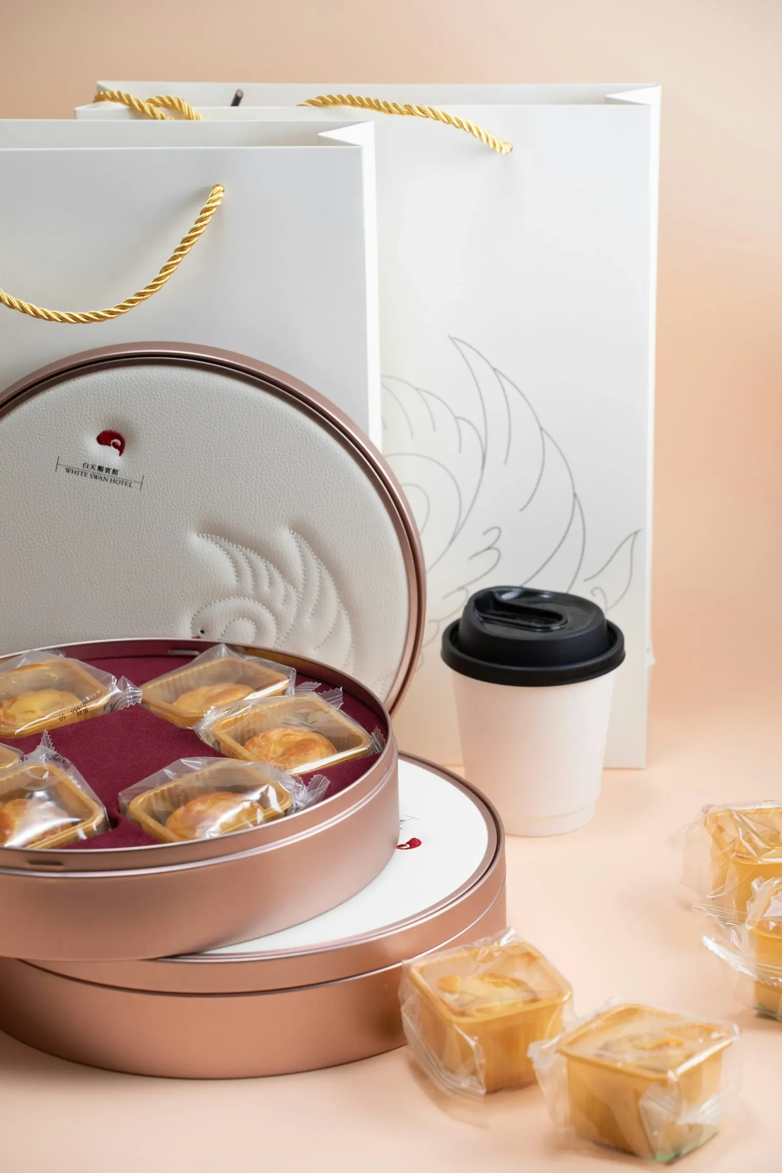 Round gift tin of pastries beside a white paper coffee cup and gift bags with gold handles