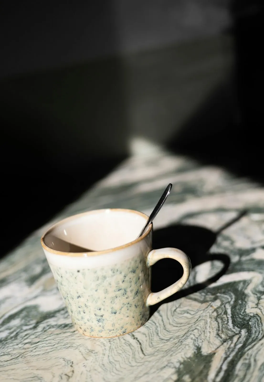 Dark speckled ceramic mug with a spoon resting inside on a green and white marble surface