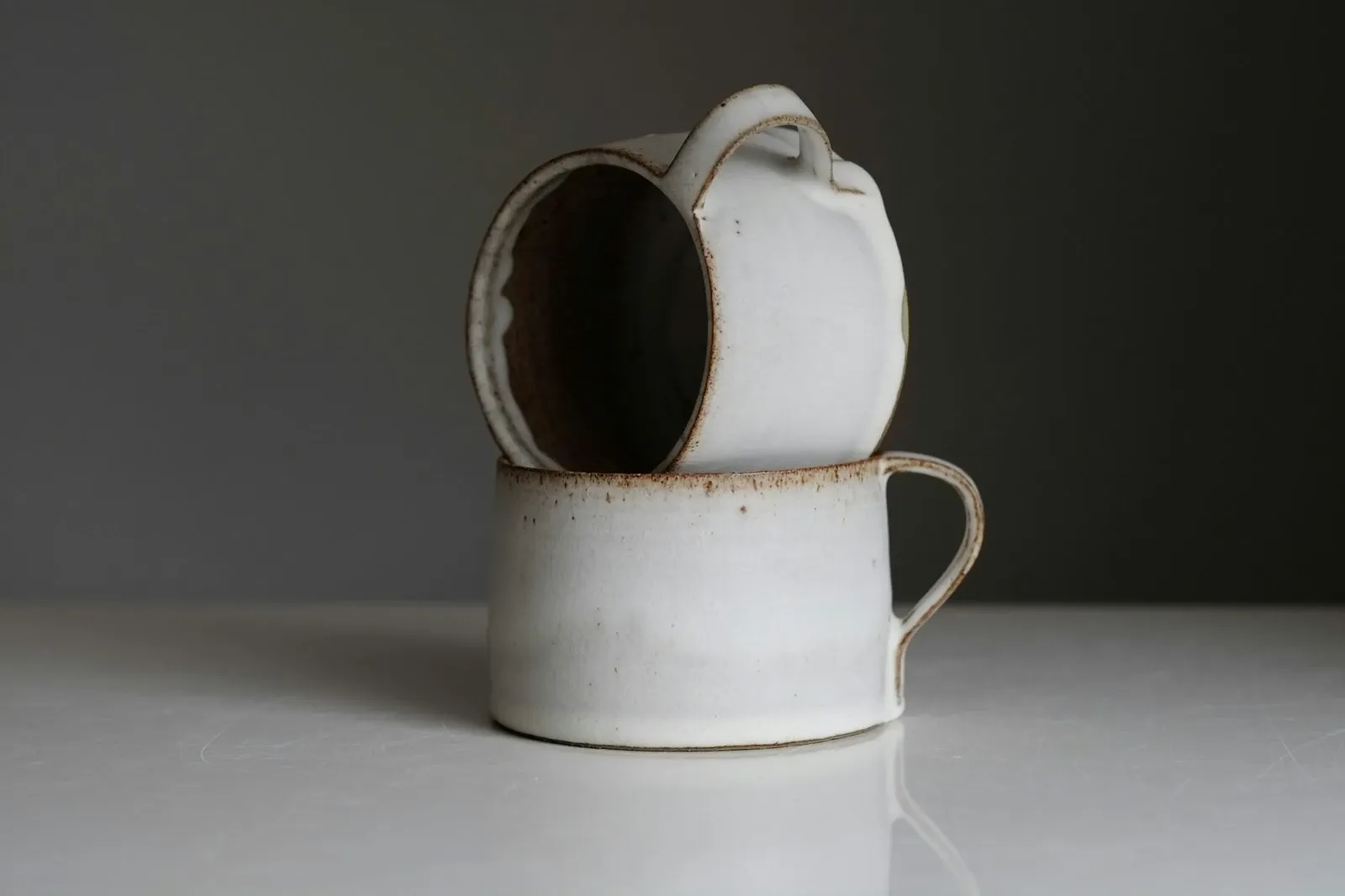 Two cream stoneware mugs stacked on a white surface against a dark gray wall
