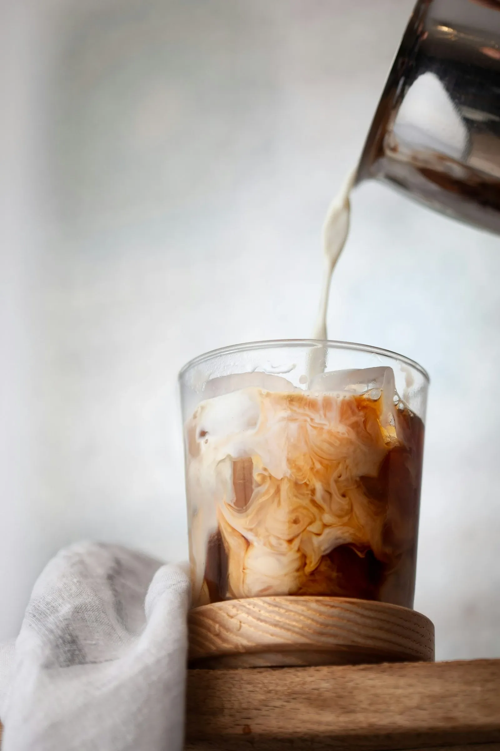 Milk pouring into a glass of iced coffee set on a round wooden coaster