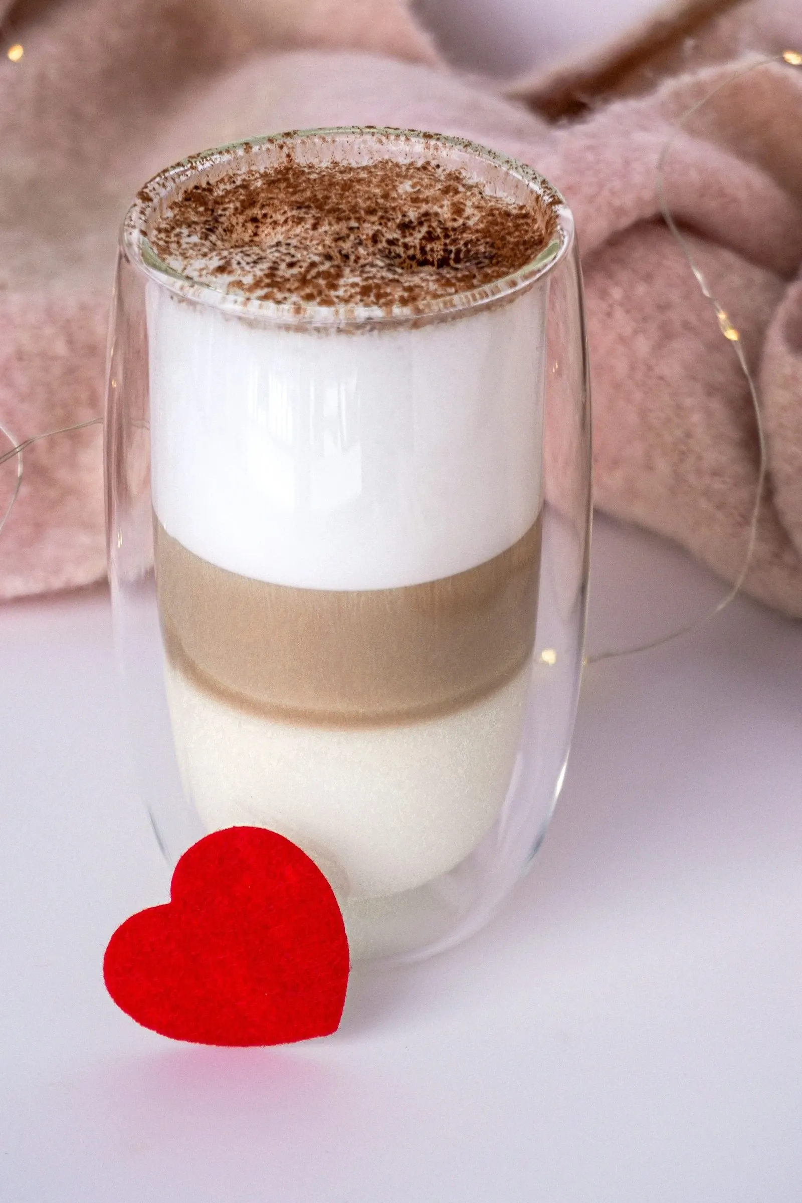 Layered latte in a double-wall glass beside a small red felt heart on a pale table